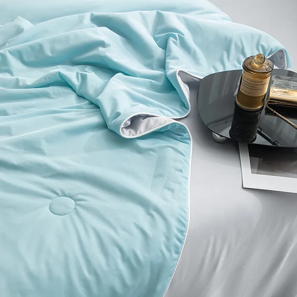 Närbild på isblått kyltäcke från NordicBlankets – svalkande material med grå kant, liggandes på vit säng med svart bricka och parfymflaska