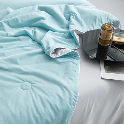 Närbild på isblått kyltäcke från NordicBlankets – svalkande material med grå kant, liggandes på vit säng med svart bricka och parfymflaska